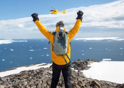 Ein Mann in einem gelben Parka steht auf einem Berg oberhalb des Meeres und reckt die Arme in die Höhe