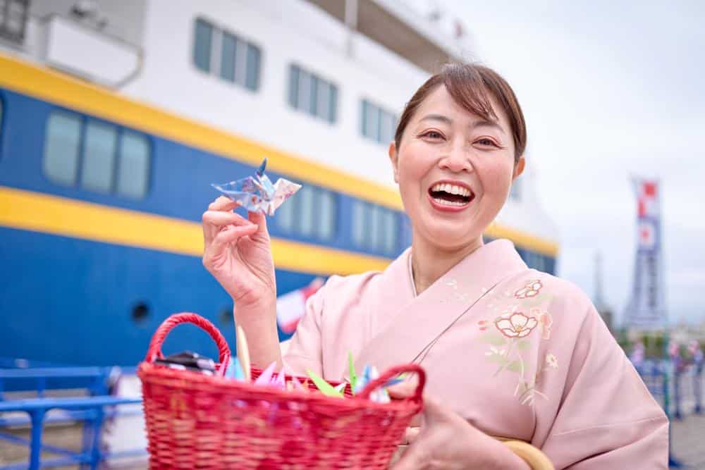 A smiling Asian woman holds an origami crane in her hands, with a large ship behind her.