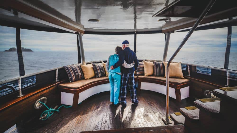 swell_achterndeck_maple-leaf-adventures Lounge auf einem Schiff mit Panoramfenster, in deren Mitte ein Paar in bequemer Kleidung den Ausblick genießt.