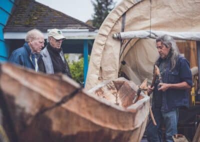 Drei Männer stehen neben einem Holzkanu, das gerade gebaut wird