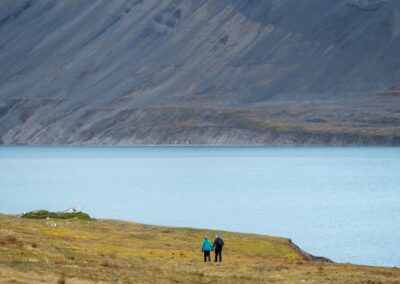 Zwei Wanderer an einem See vor einer schroffen Bergwand in Spitzbergen