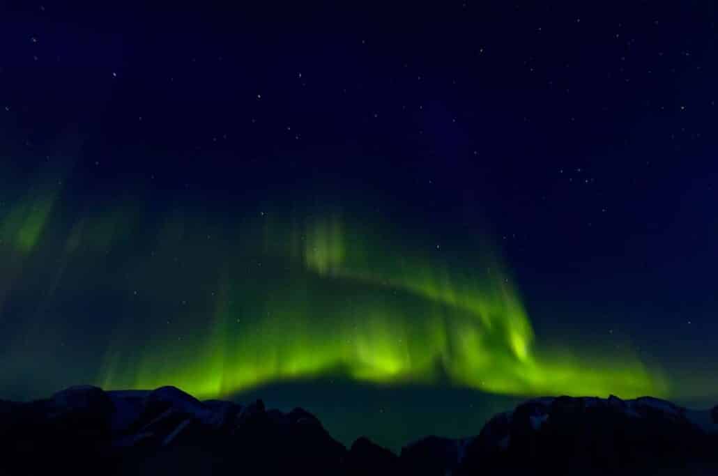 Aurora Borealis als grüner Bogen über schwarzer Bergsilhouette in Ostgrönland