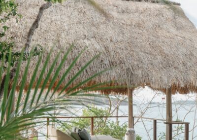 A daybed under a thatched palm roof next to the pool of a casita