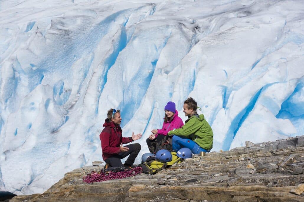 Guide und zwei Gäste sitzen mit Seil und Helmen vor der blauen Eiswand des Svartisen