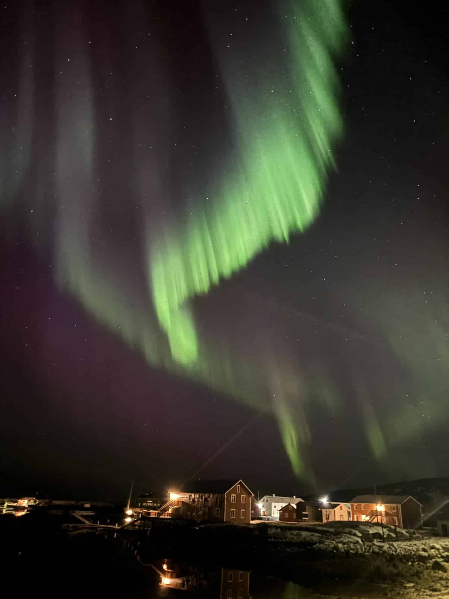 Kräftiges grünes Nordlichtband über der Insel, darunter die beleuchteten Holzhäuser am Wasser