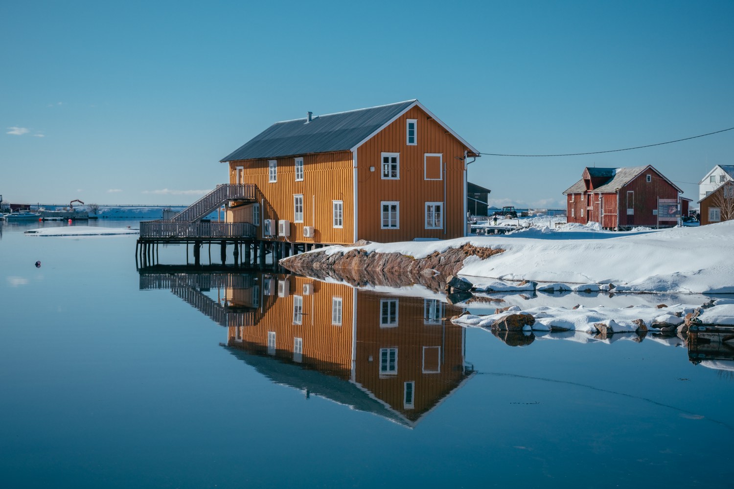 Großes ockerfarbenes Holzhaus auf Pfählen über dem Wasser mit Außentreppe, gespiegelt im windstillen Winterwasser