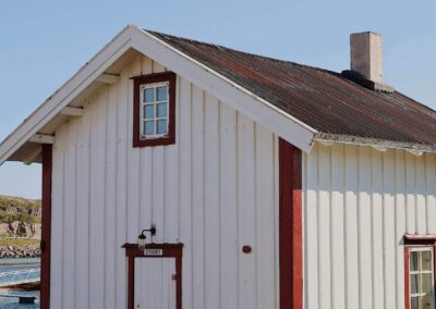 Kleines weißes Holzhaus mit roten Fensterrahmen und rostrotem Wellblechdach, über der Tür ein Emailleschild mit der Aufschrift Stabburet, dahinter der Hafen von Støtt