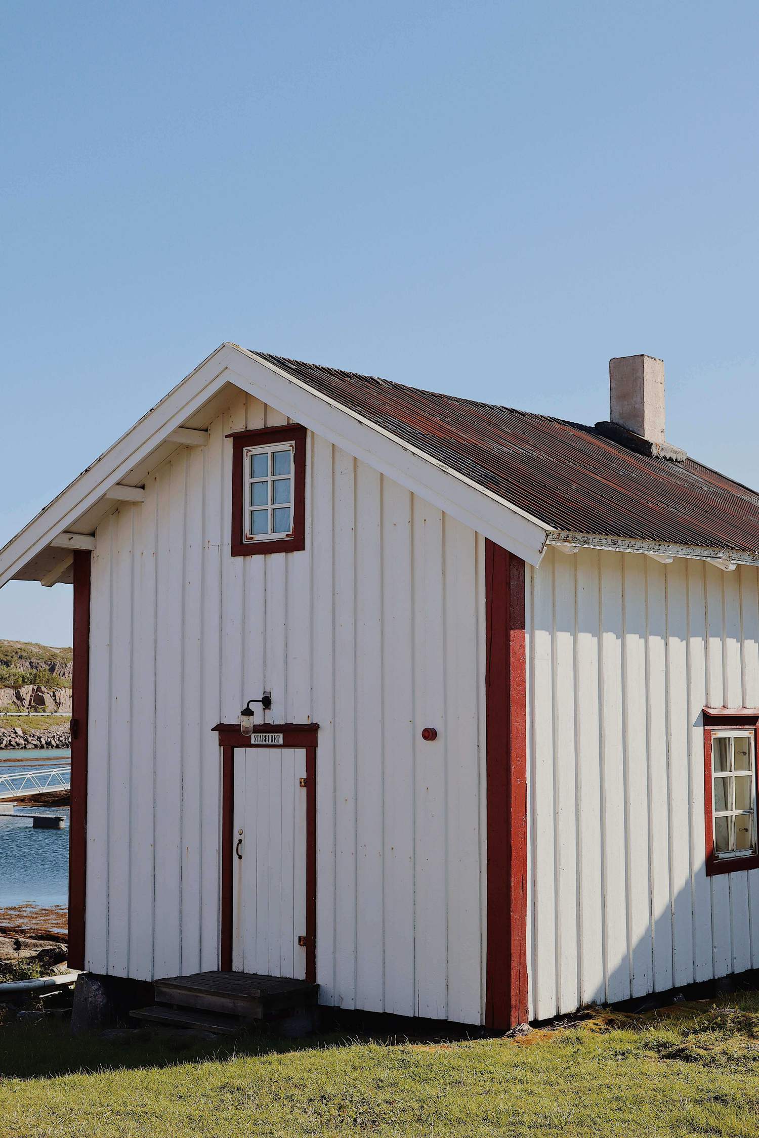 Kleines weißes Holzhaus mit roten Fensterrahmen und rostrotem Wellblechdach, über der Tür ein Emailleschild mit der Aufschrift Stabburet, dahinter der Hafen von Støtt