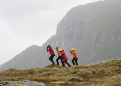 Vier Wanderer gehen gebeugt gegen den Wind über einen Grasrücken vor einer Felswand