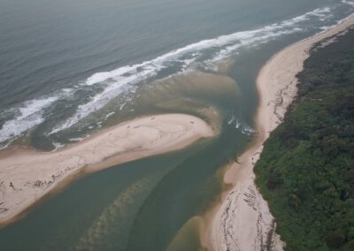 Aerial view of a river mouth flowing into the sea, featuring light-colored sandbars, surf, and strips of rainforest, Loango National Park, Gabon