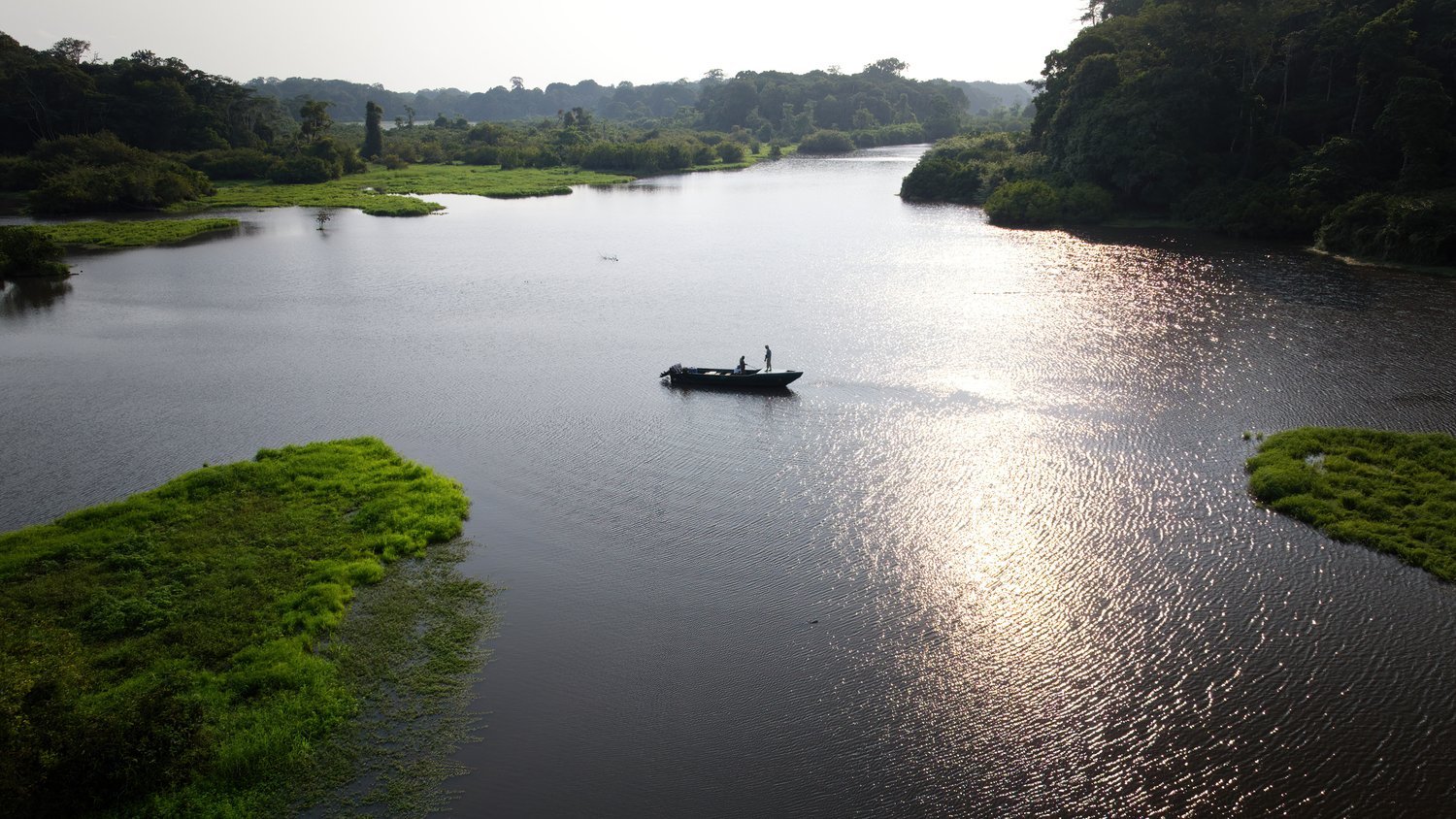 Mit dem Boot den Akaka hinab, das Abendlicht auf dem Wasser A small boat carrying two people on a wide, mirror-like river, set against the backdrop of floating islands of aquatic plants and rainforest, backlit