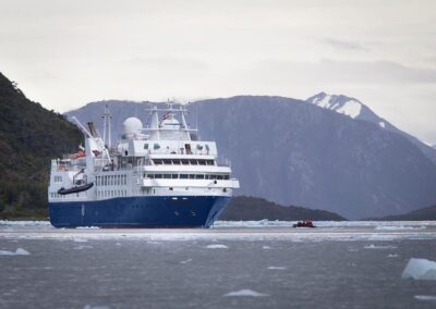 Die MS Discoverer mit dunkelblauem Rumpf vor bewaldeten Fjordbergen, ein Zodiac auf dem Wasser