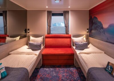 Twin cabin on the MS Hondius with two single beds, a red sofa, and a window overlooking icebergs