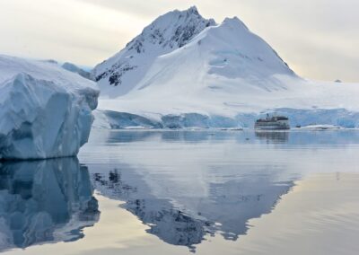 Die Greg Mortimer vor vergletscherten Bergen und einem Eisberg in der Antarktis